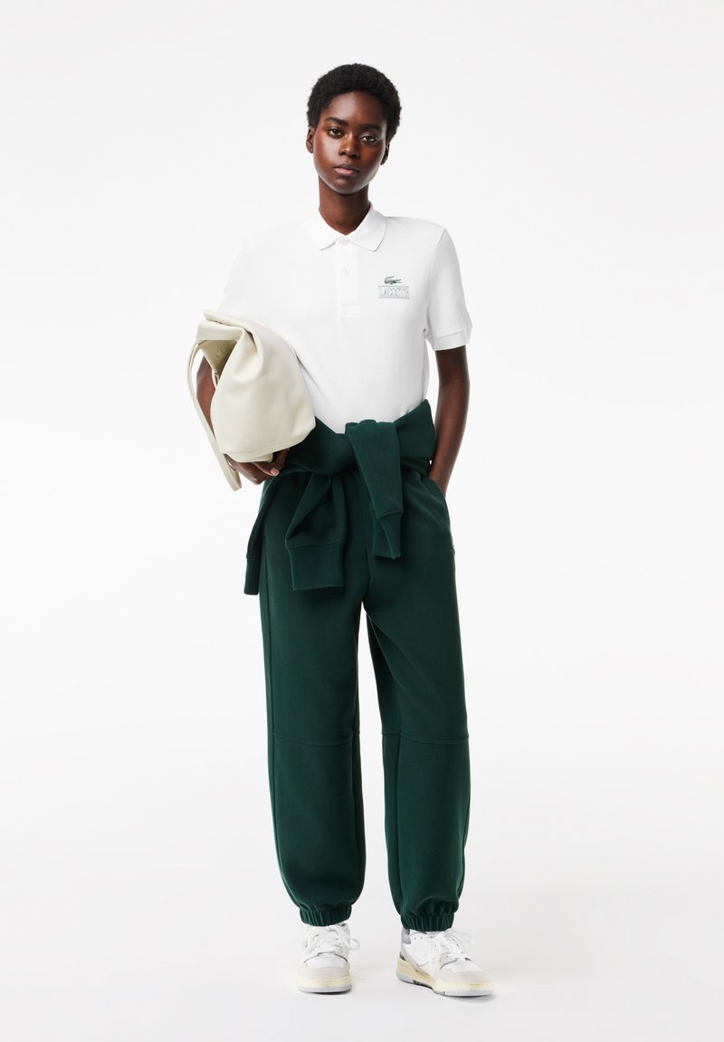 Camisa polo blanca con el logo de Lacoste, pantalones verdes con cordón y una bolsa beige. Los conjuntos presentan texturas suaves y un ajuste relajado. Zapatillas blancas.