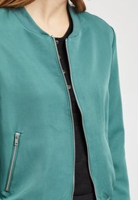 Femme portant une veste zippée bleu sarcelle par-dessus une chemise noire boutonnée, montrant le torse supérieur et le cou sur un fond uni.