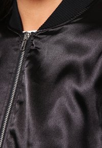 Schwarze Satin-Bomberjacke mit silbernen Reißverschluss, geripptem Kragen und glatter Textur, mit einem leicht gerafften Design an den Schultern.