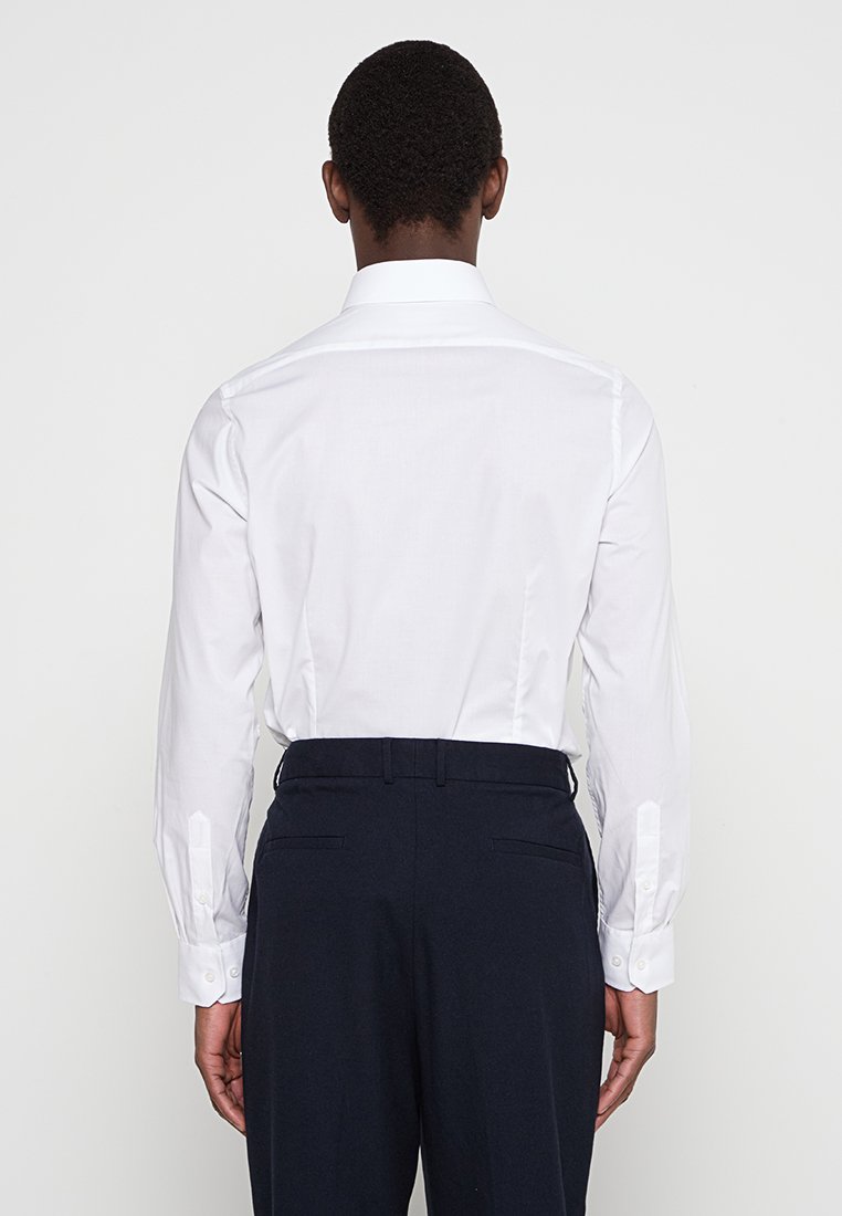 Camisa blanca de manga larga con cuello clásico, que presenta puños abotonados, combinada con pantalones oscuros de color marino. Diseño simple y limpio.