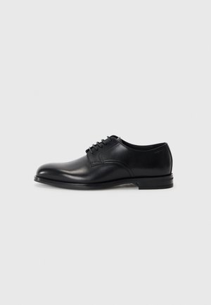 Zapato de vestir de cuero negro con punta redondeada, diseño con cordones, textura suave y suela de goma de perfil bajo. Detalles de costura mínimos.