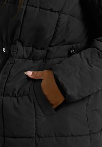 Doudoune noire avec un design matelassé, comportant une poche latérale accessible par la main. Le matériau semble lisse avec une finition mate.