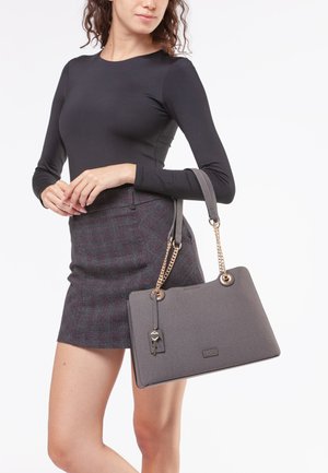 Borsa grigia testurizzata con dettagli in catena dorata e una chiave decorativa. Forma rettangolare con due manici. Indossata con un top aderente nero e una gonna a quadri.