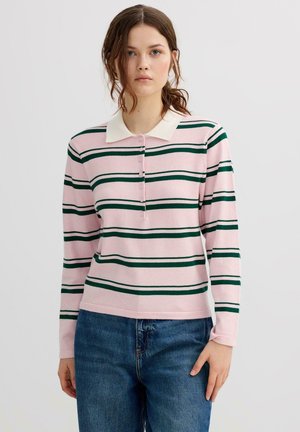 Mujer joven con una camisa polo de manga larga a rayas rosas y verde oscuro con cuello blanco y jeans azules, de pie frente a un fondo liso.