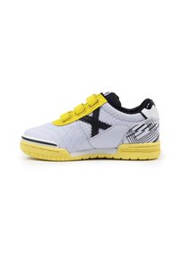 Zapatilla deportiva blanca y amarilla con diseño en negro en forma de "X", tres tiras de Velcro amarillas y un tacón con un patrón, sobre un fondo blanco.