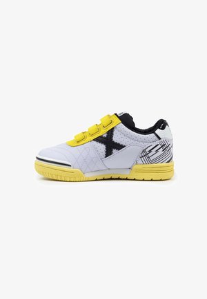Zapatilla deportiva blanca y amarilla con diseño en negro en forma de "X", tres tiras de Velcro amarillas y un tacón con un patrón, sobre un fondo blanco.