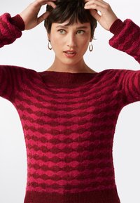 Rosa und burgunderfarbener Pullover mit einem Rautenmuster, präsentiert mit einem Bootsausschnitt und strukturiertem Stoff, ergänzt durch gerippte Bündchen.