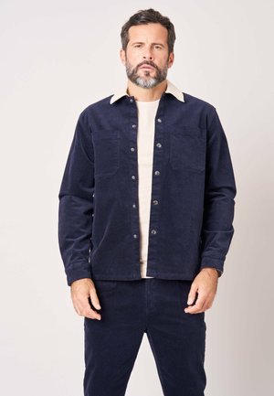 Man met baard die een donkerblauw knoopjasje met witte kraag en bijpassende donkerblauwe broek draagt, staand tegen een effen achtergrond.