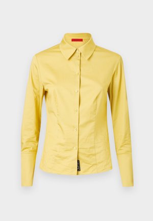 Camicia gialla a maniche lunghe con colletto a punta, in tessuto morbido, con un'etichetta nera dettagliata in basso a sinistra.