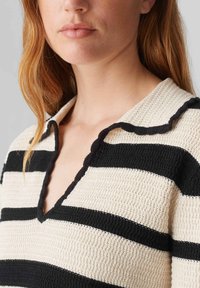 Pull en tricot à rayures crème et noires, avec un col en V et un col festonné. Tissu texturé avec un aspect confortable et épais.