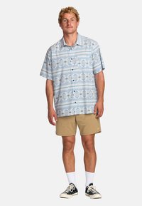 Kortærmet skjorte med knaplukning og blå geometriske mønstre på en lys baggrund, parret med beige shorts og sorte sneakers. Hvide sokker synlige.