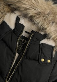 Schwarz gepolsterte Jacke mit fellummantelter Kapuze. Verfügt über einen goldenen Reißverschluss, Druckknöpfe und eine weiche weiße Innenausstattung. Strukturierter Pelz in einer hellen Farbe.