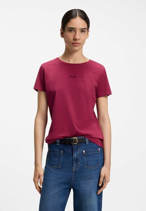 Femme aux cheveux foncés attachés en arrière, portant un t-shirt ajusté rose foncé à manches courtes et un jean bleu avec une ceinture noire sur fond blanc.