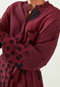 Persona che indossa un vestito bordeaux con pois neri e piccoli motivi a pois, dotato di un colletto arricciato e di una chiusura con bottoni.