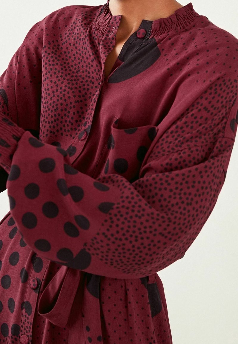 Persona che indossa un vestito bordeaux con pois neri e piccoli motivi a pois, dotato di un colletto arricciato e di una chiusura con bottoni.
