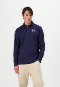 Polo de manga longa azul-marinho com colarinho preto, apresentando um logo bordado em forma de "H" em vinho. O material é suave e tem um corte clássico.