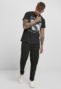 T-shirt noir avec un motif graphique et du texte, associé à un pantalon cargo noir ample. Les chaussures sont blanches, ajoutant du contraste à la tenue.