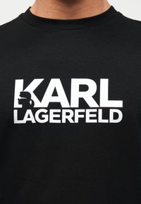 Μαύρο Τ-shirt με έντονο λευκό κείμενο "KARL LAGERFELD" και μια μικρή σιλουέτα άνδρα με γυαλιά ηλίου και αλογοουρά ενσωματωμένη στο γράμμα K.