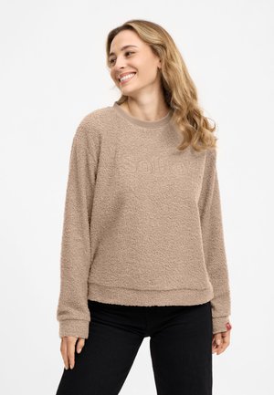 Woman smiling and looking to the side wearing a beige textured long-sleeve sweater with "Sofie" embossed and black pants.