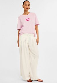 Ljusrosa bomulls t-shirt med "mon chéri" i fet röd text, kombinerad med löst sittande vita veckade byxor och minimalistiska röda sandaler.