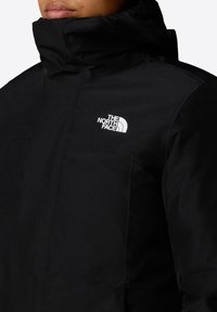 Zwarte geïsoleerde jas met hoge kraag; heeft het witte logo van The North Face. Glad materiaal en functionele ritsen op de zakken.