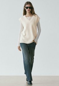 Femme portant un gilet en maille écru surdimensionné par-dessus une chemise blanche à manches longues, un jean large et foncé, des chaussures noires, et de grandes lunettes de soleil noires, marchant vers l'avant.