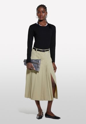 Donna con top nero a manica lunga, gonna midi plissettata beige con spacco laterale, ballerine nere, tiene una pochette argentata.