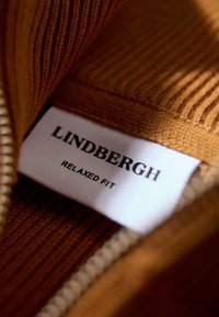Tessuto lavorato a maglia marrone con texture a coste e etichetta bianca che recita "LINDBERGH RELAXED FIT." Dettaglio con zip visibile lungo un bordo.