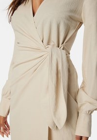 Beige omlottklänning med långa ärmar, rynkdetalj vid midjan och en knuten accent på ena sidan. Slät, mjuk tygtextur.