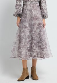 Mujer vistiendo una falda midi floral transparente y una blusa a juego con mangas abullonadas, combinada con botas de ante beige hasta el tobillo, frente a un fondo liso.