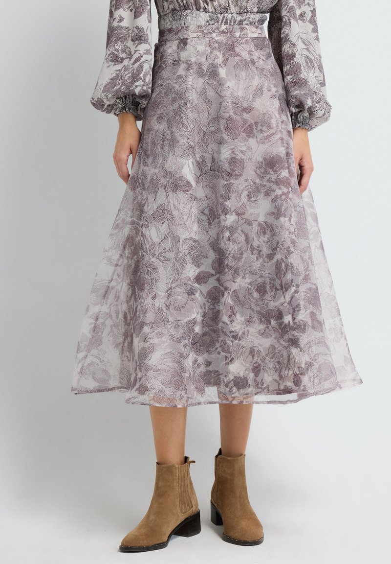 Mujer vistiendo una falda midi floral transparente y una blusa a juego con mangas abullonadas, combinada con botas de ante beige hasta el tobillo, frente a un fondo liso.