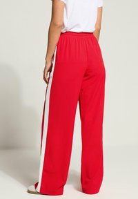 Rote weit geschnittene Hose aus weichem Stoff, mit elastischem Bund und weißen Seitenstreifen. Das Design ist lässig und bequem.