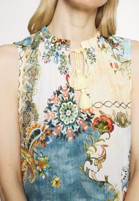 Blouse sans manches à motifs floraux en tissu léger avec des tons bleu et crème. Présente un col froncé et des accents de breloques jaunes.