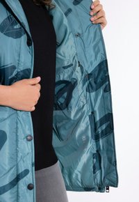Leichte türkisfarbene Pufferjacke mit schwarzem abstraktem Muster; Innentasche mit Reißverschluss; glänzende, glatte Textur; Druckknopfverschlüsse.