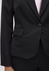 Svart blazer laget av glatt stoff, med enkelt knappelukking, klaiplommer og noterte revers. Slim fit med skreddersydd design.