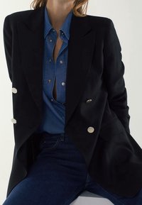 Blazer noir à double boutonnage avec des accents de boutons argentés, superposé sur une chemise en denim bleu. Look décontracté complété par un jean foncé.