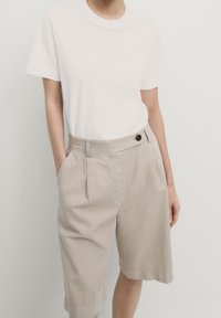 Hvid kortærmet t-shirt parret med beige cropped shorts, der har en sidelomme og en knapdetalje ved taljen. Glat stofstruktur.