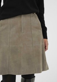 Wildlederrock im A-Linien-Schnitt in gedämpftem Grün-Grau, mit Fronttaschen, Nahtdetails und glatter Textur. Kombiniert mit schwarzen Leggings.