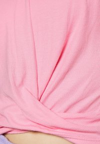 Haut en tissu côtelé rose clair avec un détail noué à la taille, présentant une texture lisse et un éclat subtil.