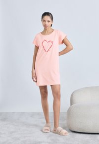 Ροζ βαμβακερό νυχτικό με κοντά μανίκια, με κόκκινο κείμενο σε σχήμα καρδιάς. Συνδυάζεται με μπεζ χνουδωτές παντόφλες σε υφασμένο γκρι χαλί.