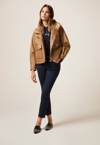 Veste en cuir synthétique marron avec un design court, deux poches avant et des fermetures à bouton-pression. Portée sur un haut bleu marine et associée à un pantalon sombre.