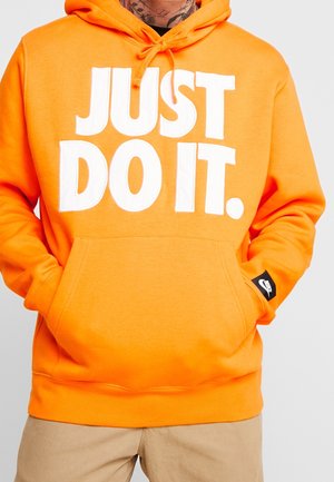 Osoba ubrana w pomarańczową bluzę z kapturem z białym napisem "JUST DO IT." i ręce w przedniej kieszeni, widoczne beżowe spodnie.