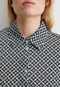 Kragenbluse mit einem geometrischen Muster in Schwarz, Weiß und Türkis, mit Knopfverschlüssen und einer glatten Stoffoberfläche.