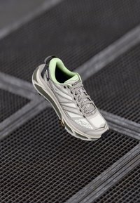 Light grey running shoe with mesh upper and green accents. Features a thick sole, laces, and a black heel tab. Vibram logo on sole.