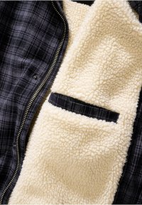 Melna un pelēka pārklājoša kārta ar zelta rāvējslēdzēju; balta vilnas oderes iekļauj mazu kabatu ar melnu akcentu. Mīksta tekstūra visā virsmā.