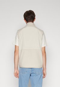 Beige fleecevest med høy krage og paneldesign, kombinert med en lett hvit t-skjorte med korte ermer og lyseblå jeans.