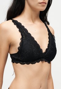 Γυναίκα που φοράει μαύρο δαντελένιο bralette με ανθισμένα σχέδια και κυματιστά άκρα, εμφανιζόμενη από τους ώμους έως την μέση σε απλό φόντο.