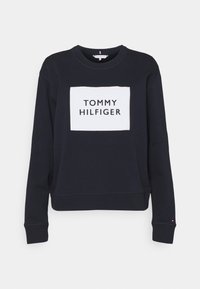 Felpa girocollo blu navy con maniche lunghe e una patch rettangolare bianca sul petto che riporta la scritta "TOMMY HILFIGER" in lettere nere.