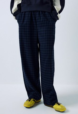 Person wearing loose dark blue checkered pants, a black and white jacket, and bright yellow sneakers standing against a plain background.