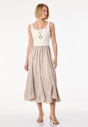 Vrouw draagt een mouwloos wit topje, een beige gestructureerde midi-rok, een ketting met hanger en beige sandalen met hak, staande tegen een witte achtergrond.